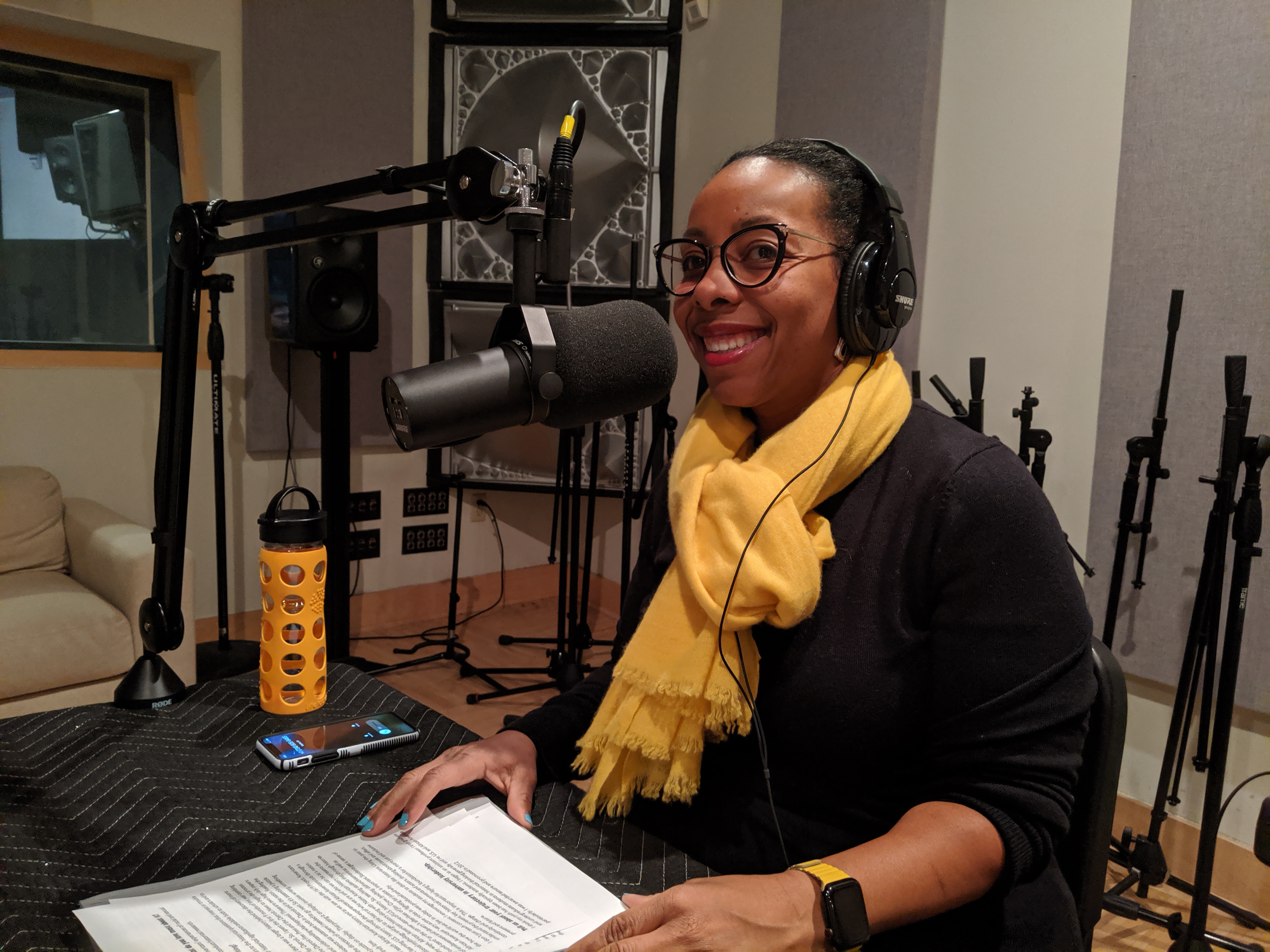 Kidada Williams in front of a microphone wearing headphones, black shirt and yellow scarf