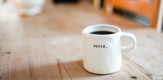 a hot beverage in a white ceramic mug with the word BEGIN sits on a wooden table