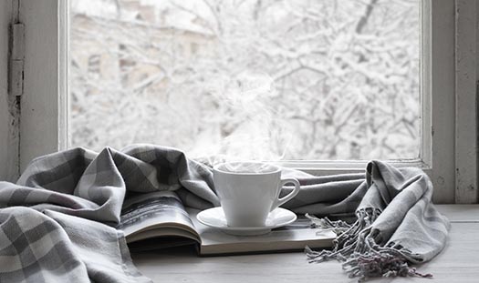 a cup of warm beverage steams on a windowsill near a book and blanket - outside the window the world is covered in snow