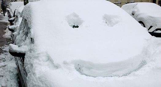a line of cars parked on a snowy street, under a significant amount of snow, the front car has a smile drawn in the snow on the windshield